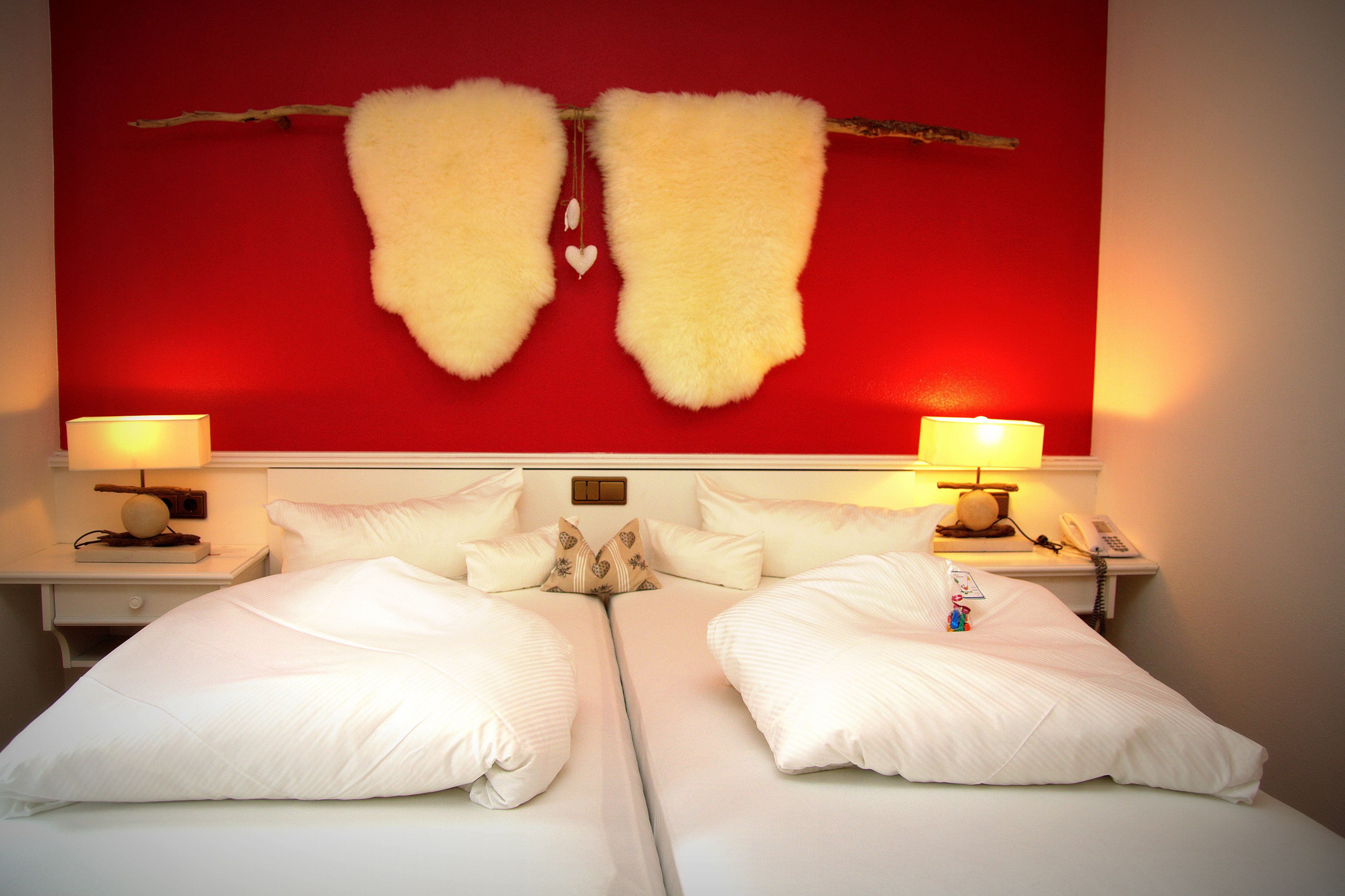 Habitación de hotel con dos camas, ropa de cama blanca, pared roja decorada con una rama y un corazón, y lámparas de noche.