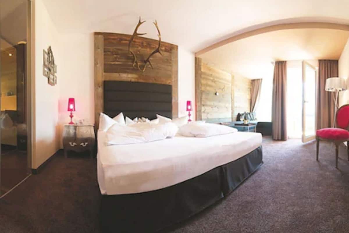 Habitación de hotel con una cama grande, cabecera de madera, un separador de vidrio, una silla roja y una mesita de noche con lámpara.