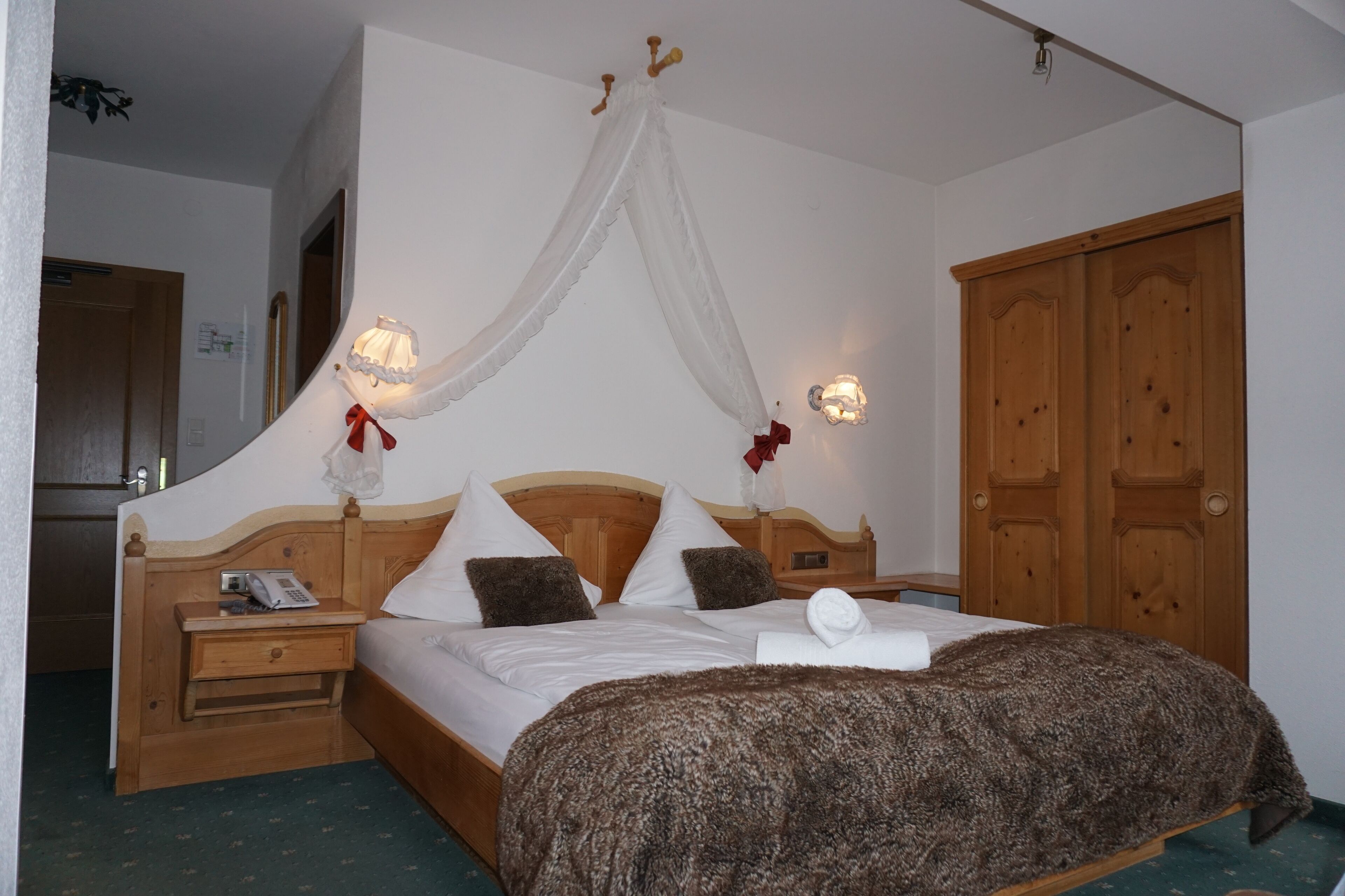 A bedroom with a wooden bed, a nightstand with a phone, a bedside table, a wardrobe, and a canopy bed with a red ribbon.