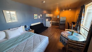 Quarto de hotel pequeno, com cama, mesa de cabeceira, uma kitchenette, mesa de jantar com cadeiras e janela com cortinas.
