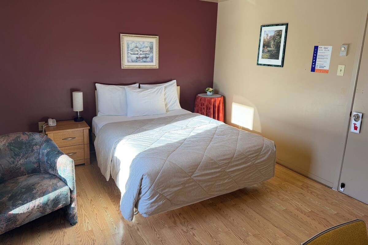A bedroom with a bed, a nightstand, a lamp, a chair, and a small table with a plant.