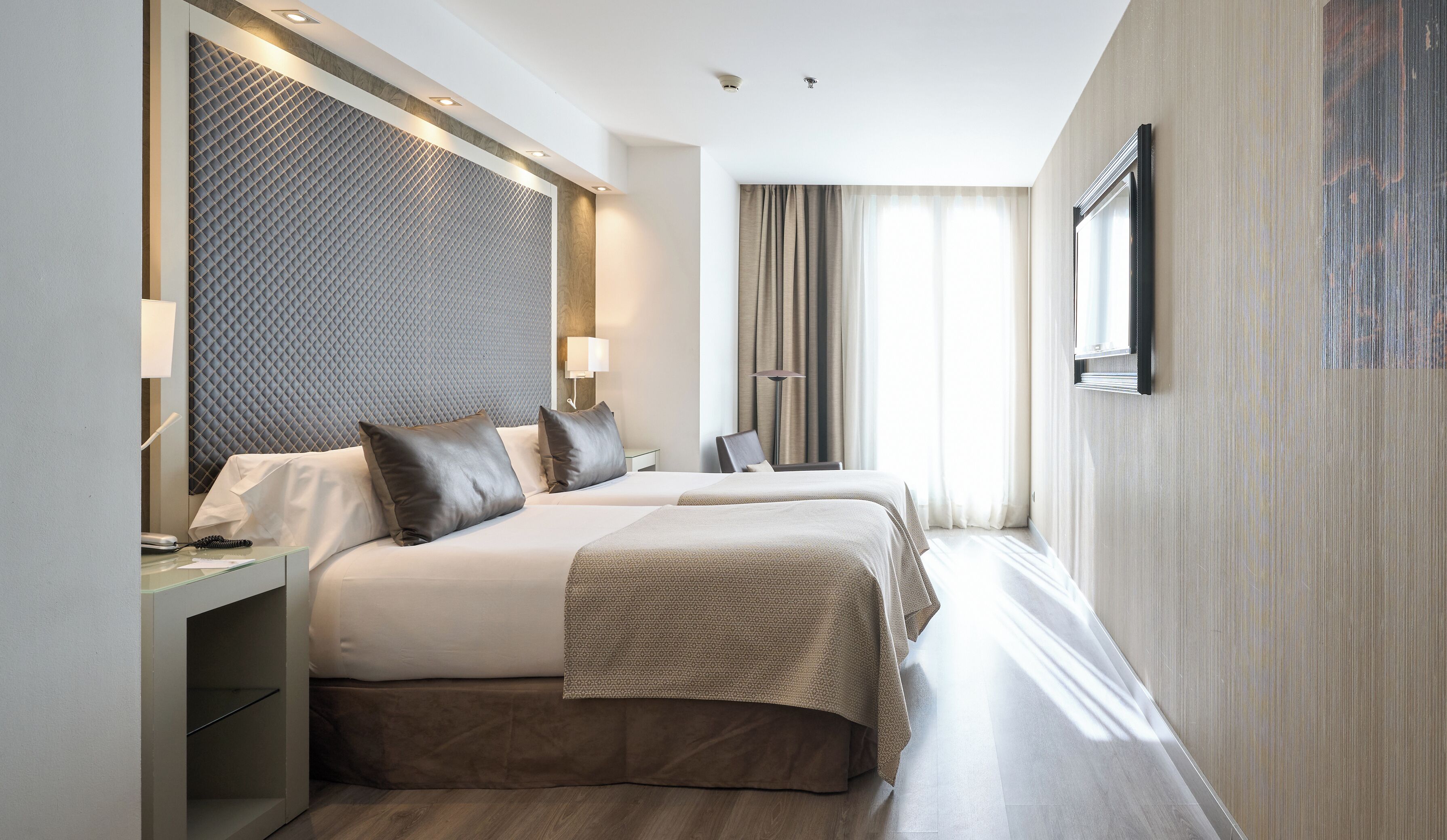 Une chambre d’hôtel avec deux lits, un bureau, une lampe, une fenêtre avec des rideaux et un téléviseur fixé au mur.