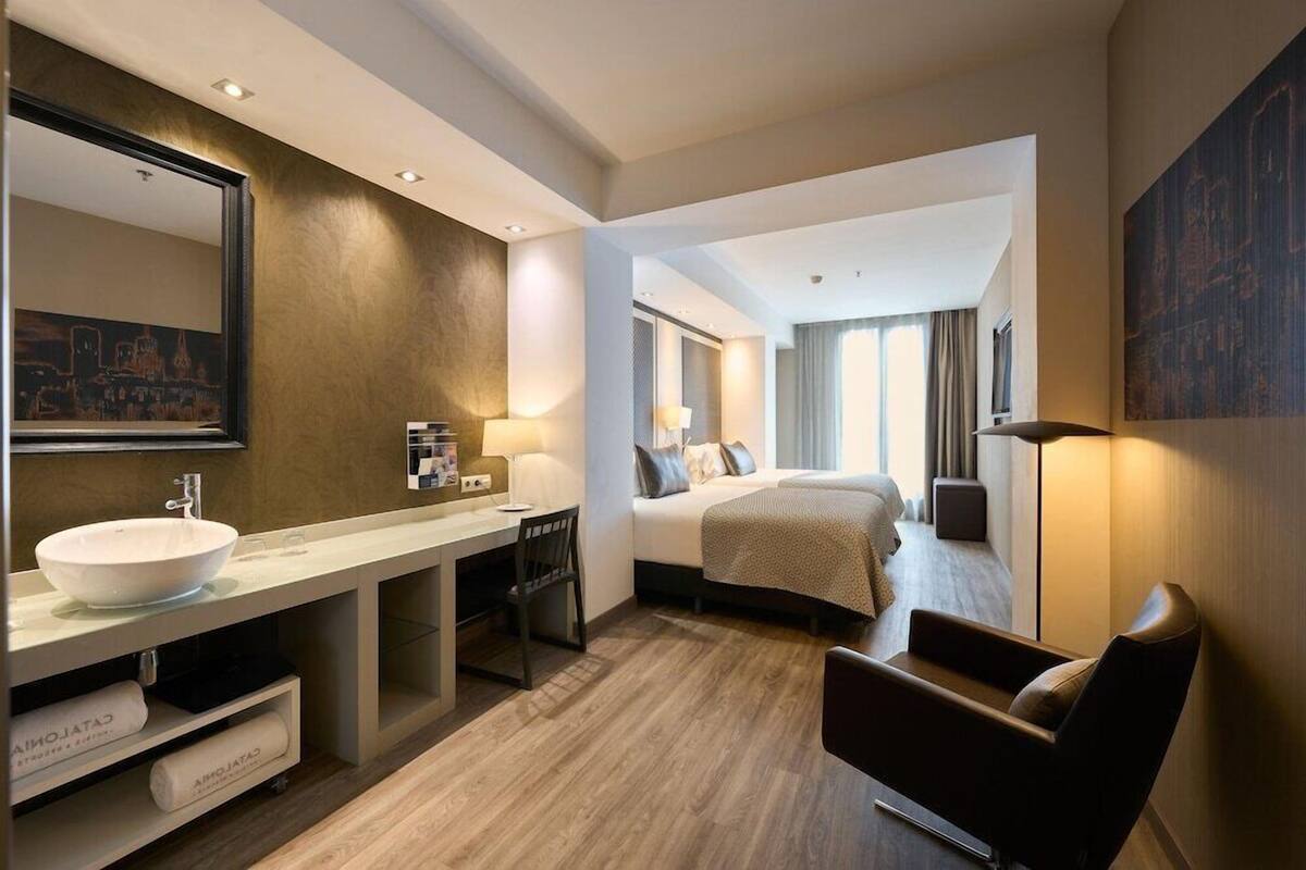 A hotel room with a large mirror, a sink, a bed, a chair, and a desk.