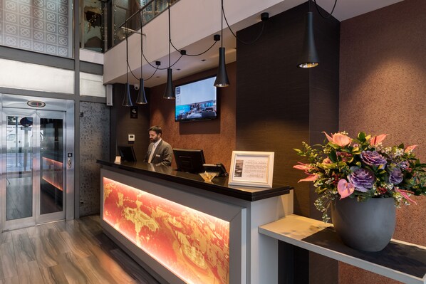 A hotel reception area with a modern design, including a reception desk, a large screen displaying a cityscape, and a potted plant with flowers. - Catalonia Square (Barcelona)