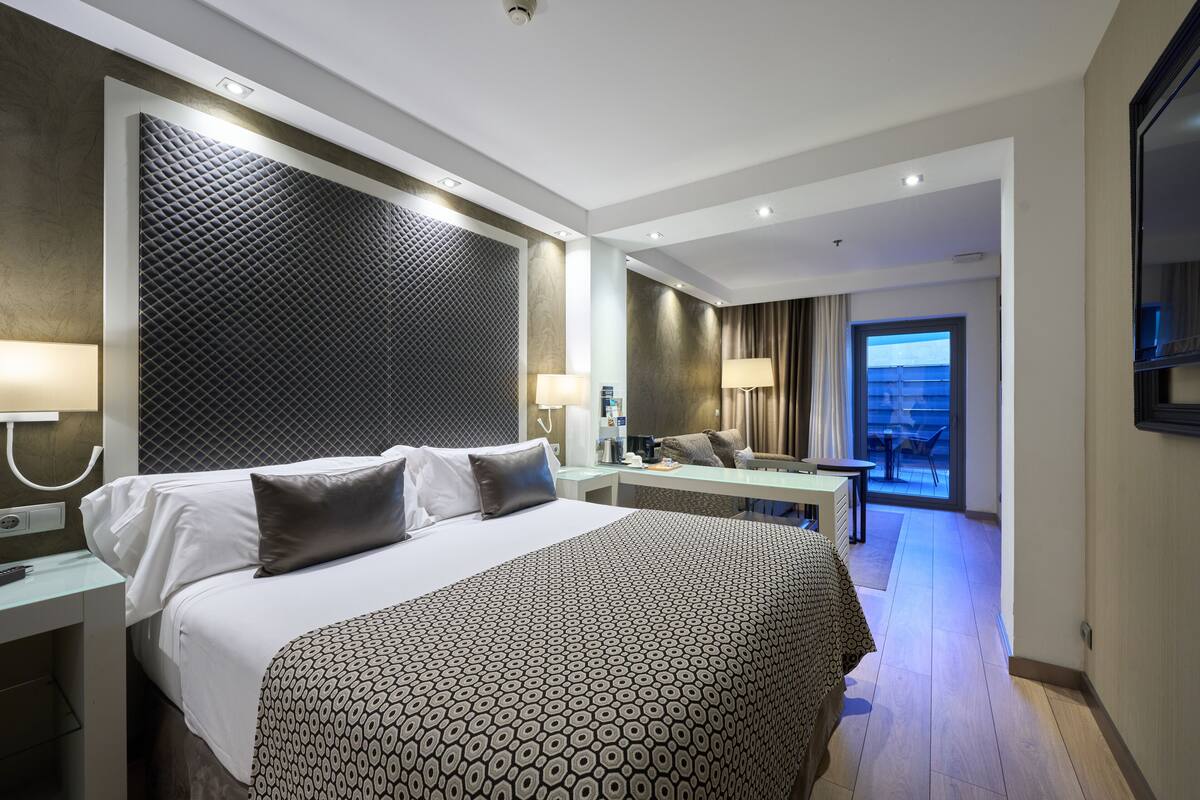 A modern hotel room with a large bed, a desk, and a view of the outdoors.