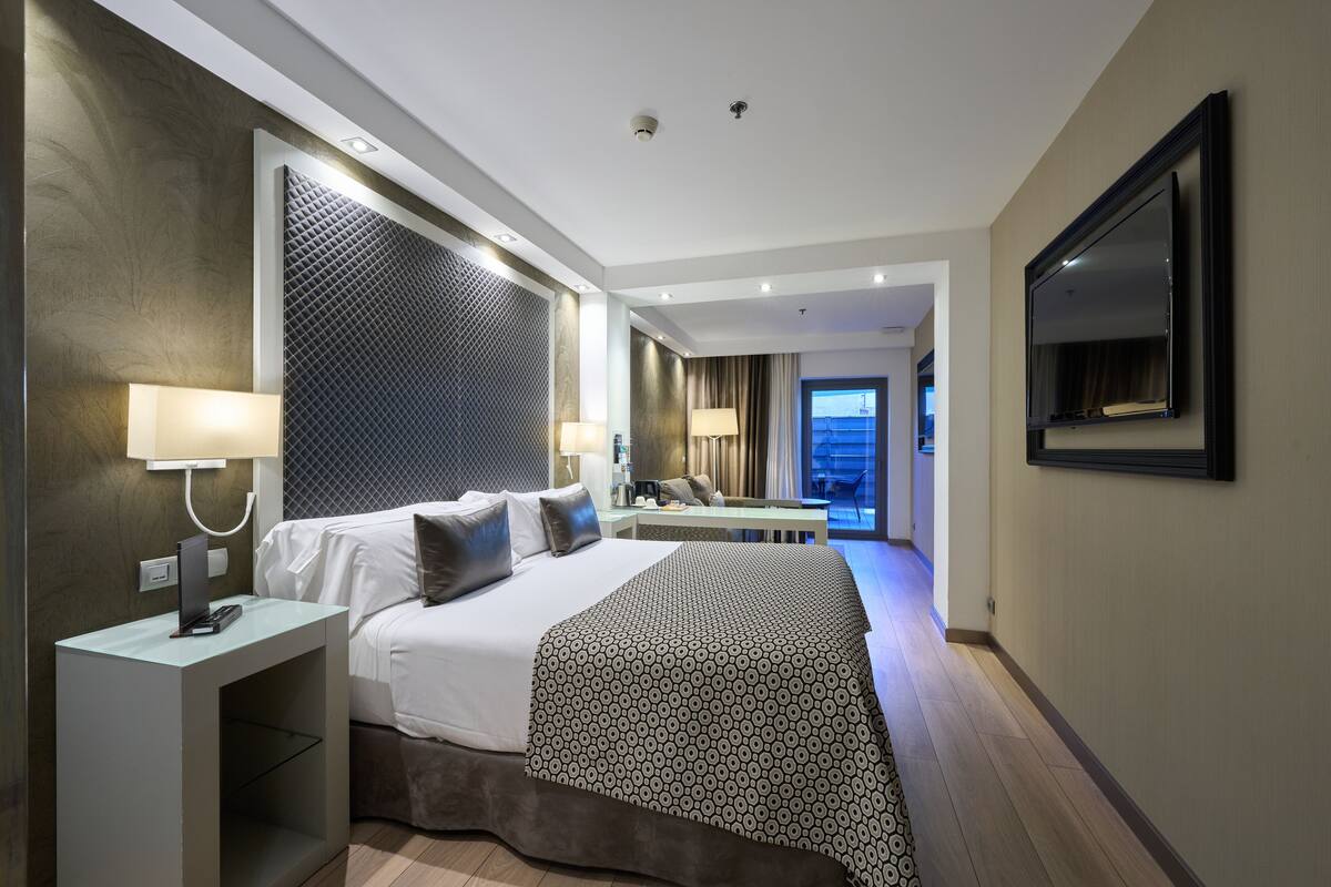 A modern hotel room with a large bed, a desk, a TV, and a view of the city.
