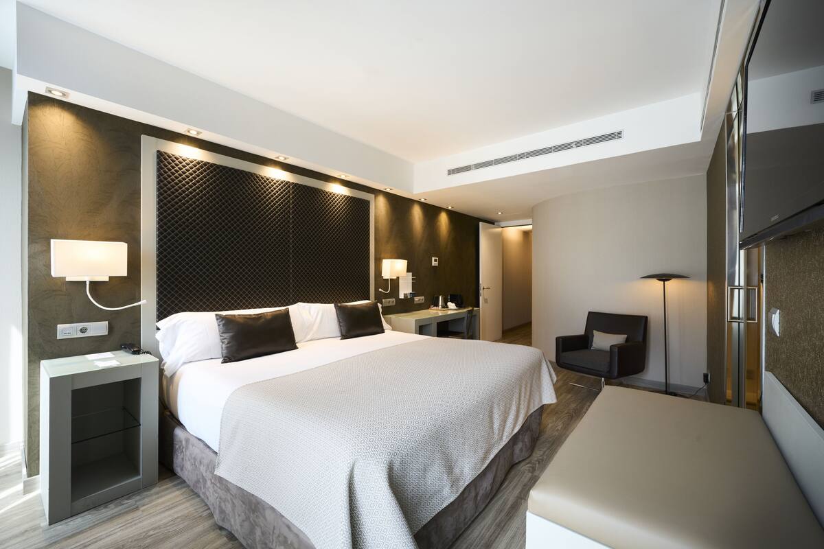 A modern hotel room with a large bed, bedside tables, a desk, a chair, and a wardrobe.