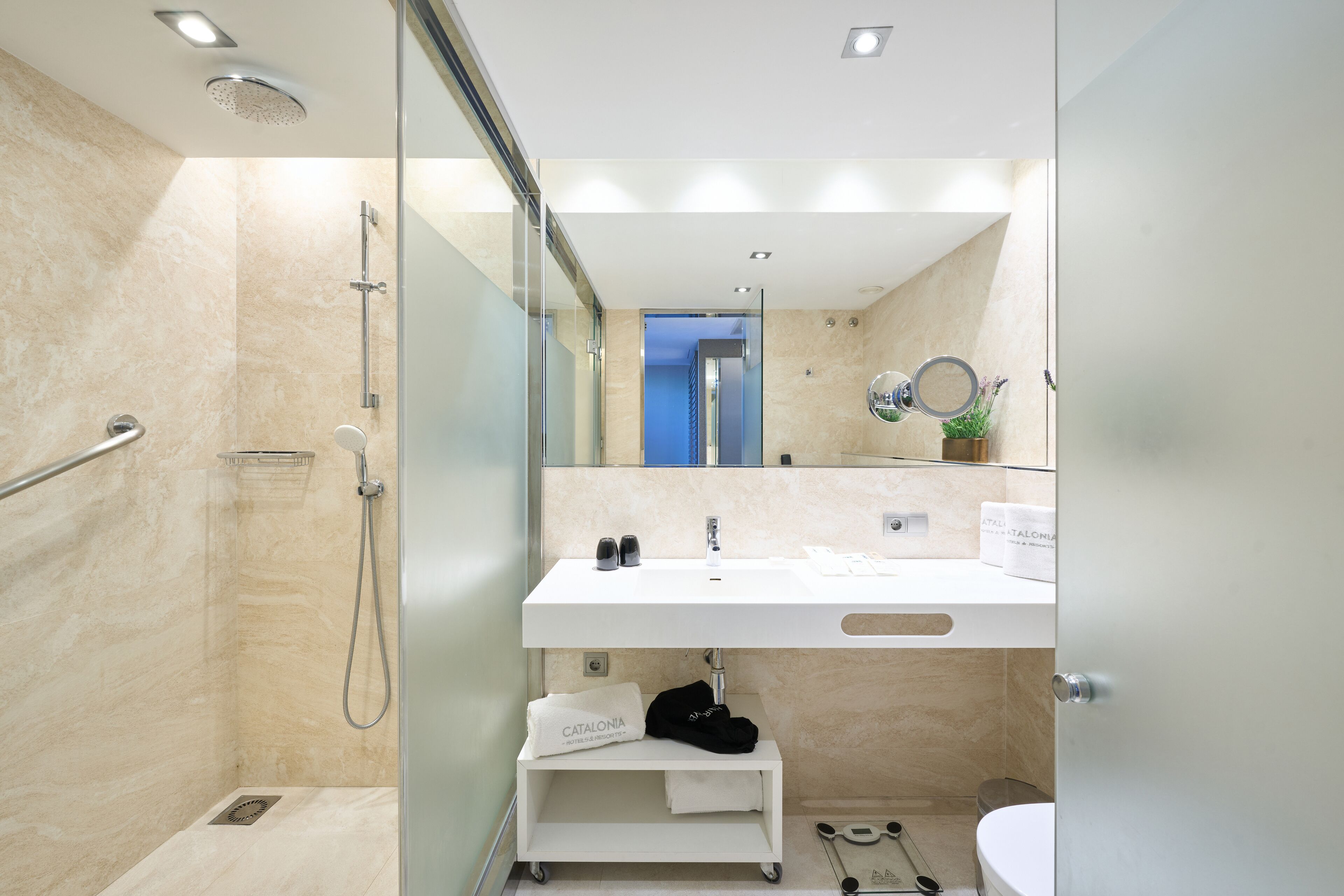 A modern bathroom with a glass-enclosed shower, a white sink with a mirror above, and a small shelf with toiletries.