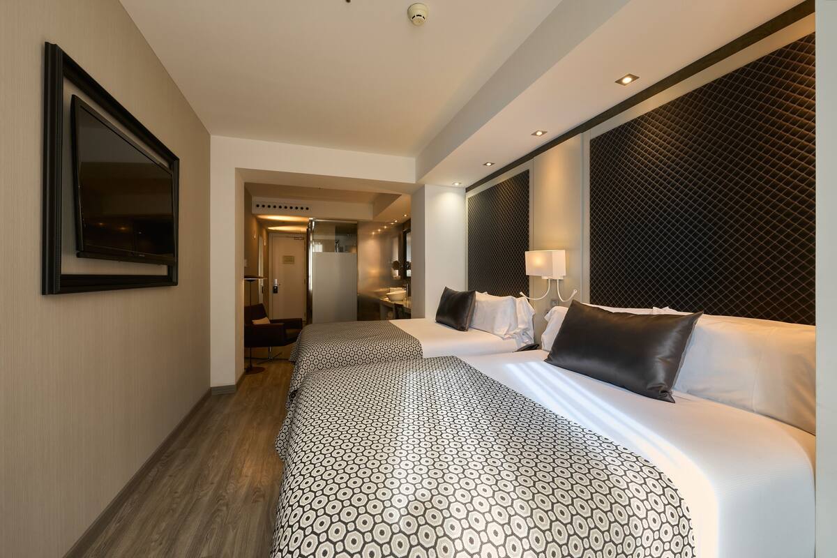 A hotel room with two beds, a large mirror, a flat-screen TV, a modern headboard, and a hallway leading to a bathroom and kitchen area.