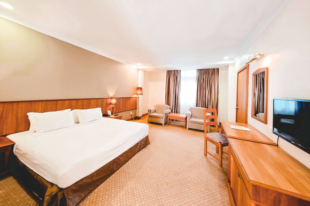 Ein Hotelzimmer mit einem großen Bett, zwei Sesseln, einem Holztisch, einem Flachbildfernseher und einem großen Fenster mit Vorhängen.