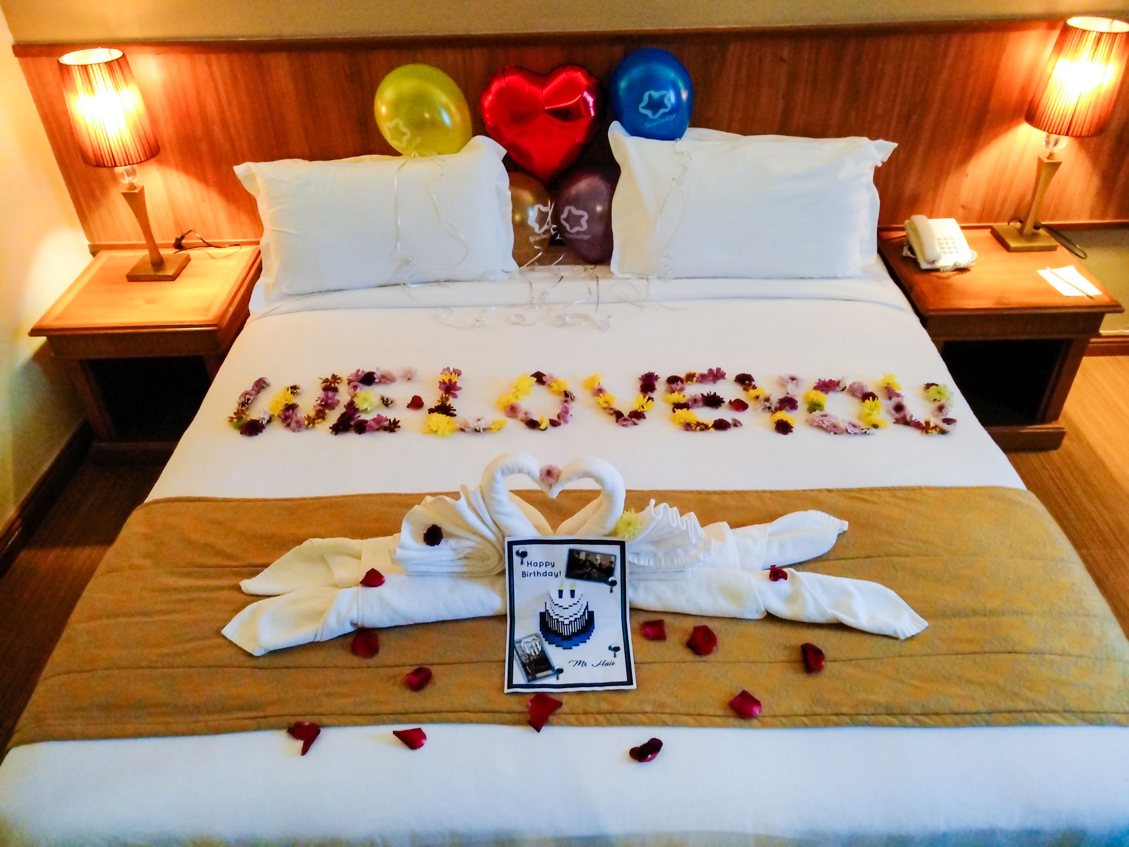 Habitación de hotel con una cama decorada con la palabra 'LOVE' hecha de flores, un arreglo de toallas con forma de cisne y una tarjeta de cumpleaños.