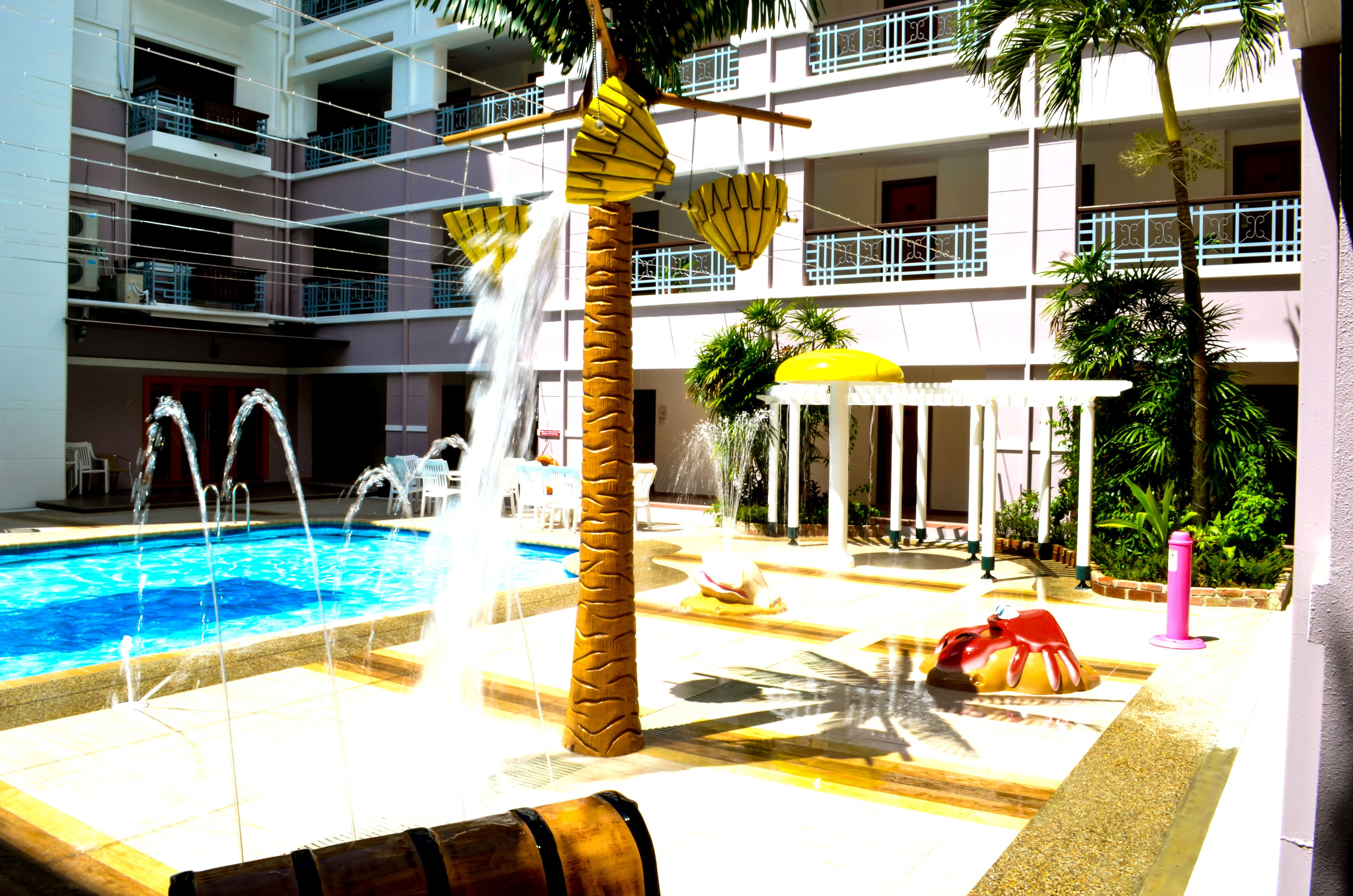 A pool area with multiple water features, including a central fountain and water jets, surrounded by palm trees and a covered seating area.