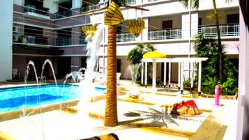 A pool area with multiple water features, including a central fountain and water jets, surrounded by palm trees and a covered seating area.