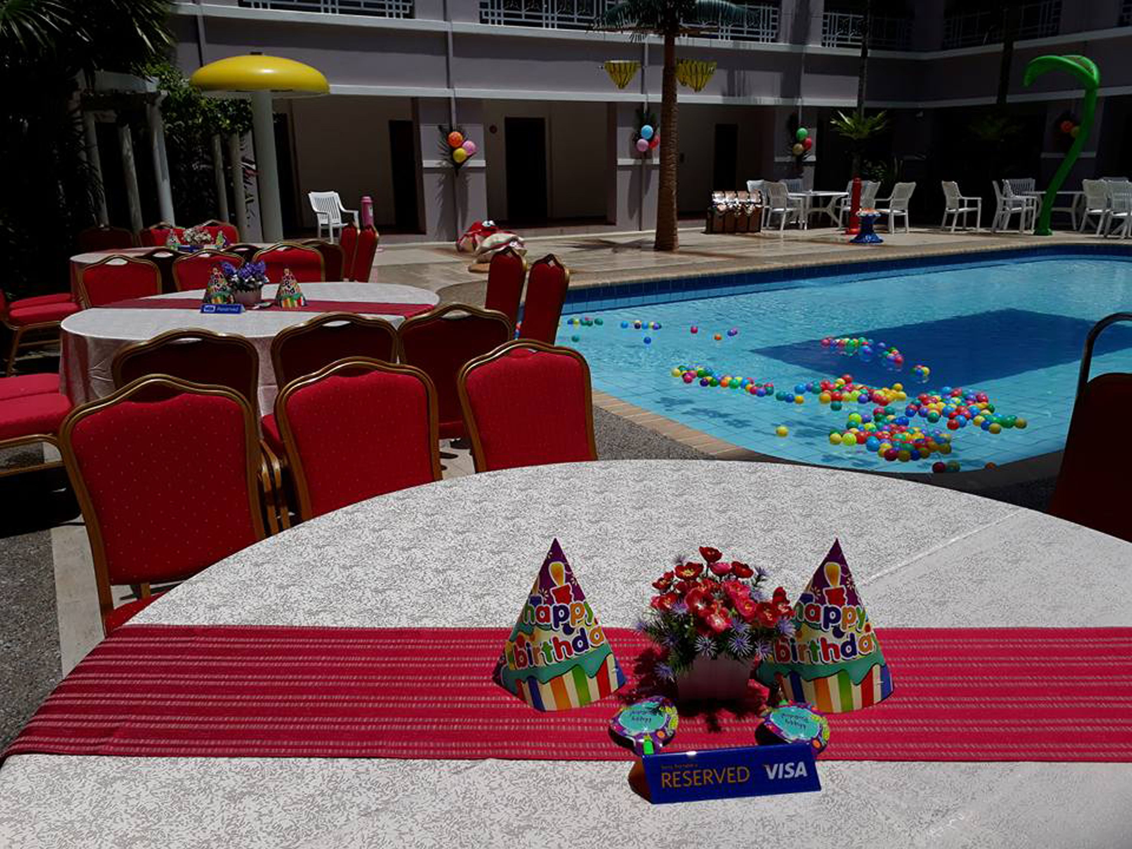 Zona de comedor junto a la piscina con sillas y mesas rojas, una piscina con pelotas de colores y un letrero de 'Reservado para VISA'.