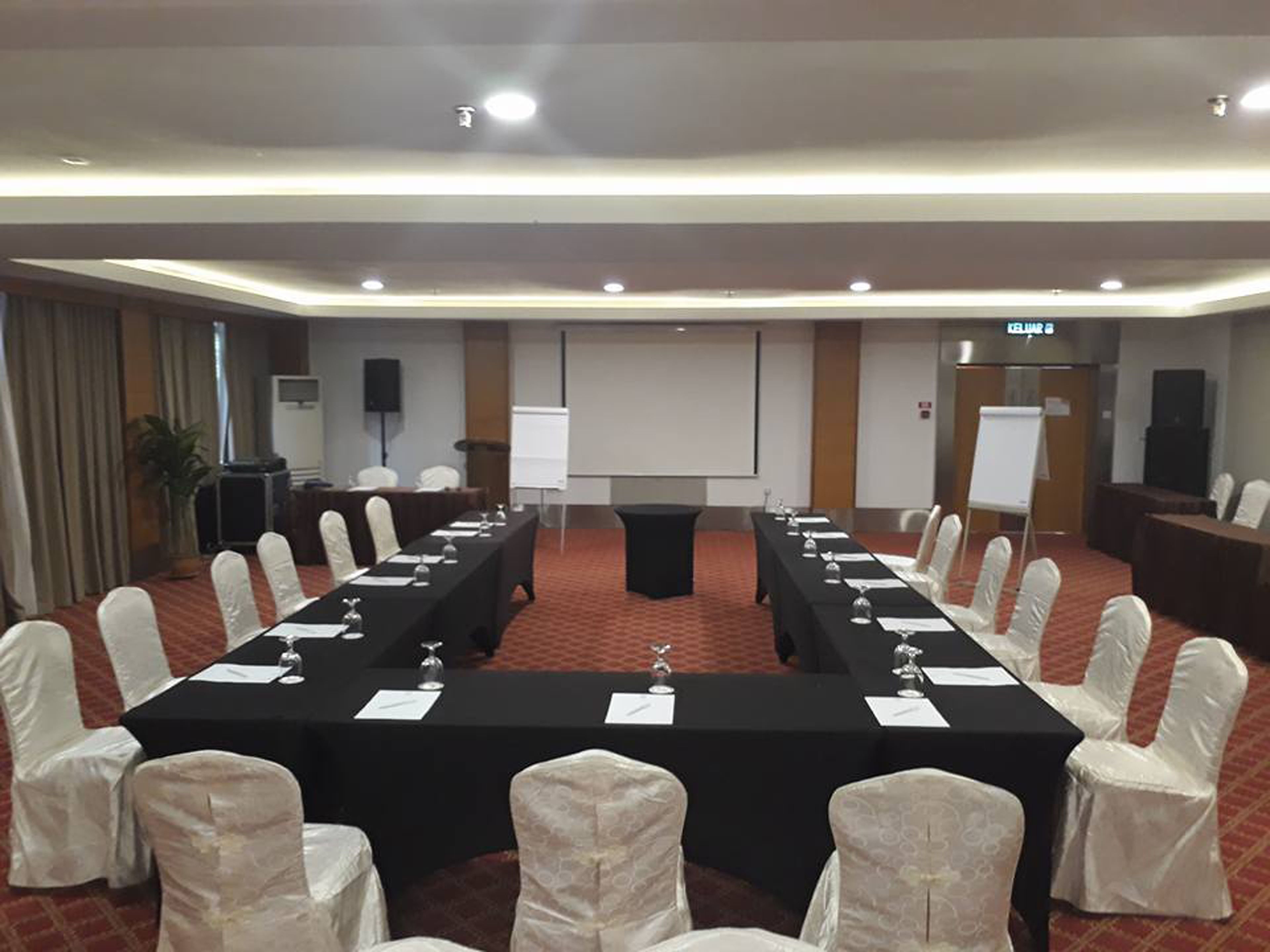 Un salón de conferencias con una larga mesa, sillas blancas, una pantalla de proyector y una pizarra.