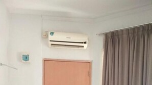 Una habitación con una puerta de madera cerrada, un equipo de aire acondicionado en la pared, un bote de basura rojo y una cortina al lado derecho.