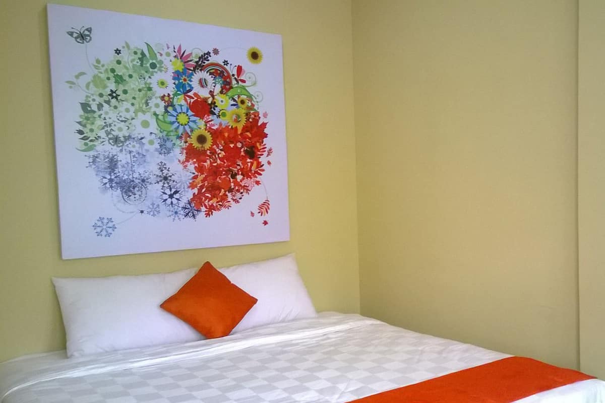 Een bed met wit en oranje beddengoed, een kleurrijk abstract schilderij aan de muur, en een enkel oranje kussen.
