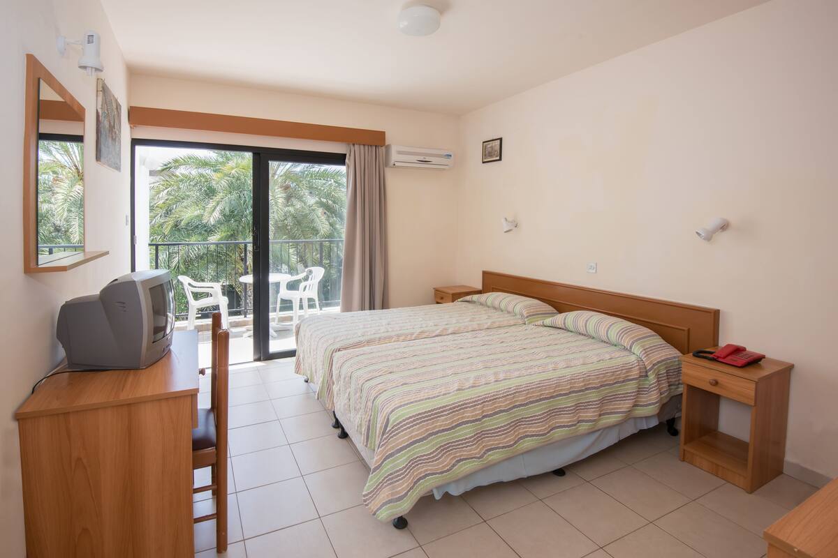 Ein Schlafzimmer mit einem Bett, einem Schreibtisch mit Fernseher, einem Sessel, einem Nachttisch und einem Balkon mit Blick auf Palmen.
