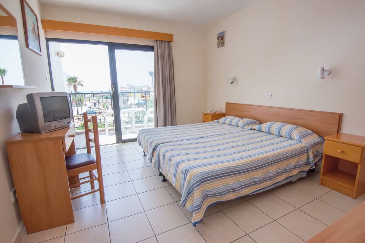 Ein Hotelzimmer mit einem Bett, einem Schreibtisch mit Fernseher, einem Sessel, einem Nachttisch und einem Balkon mit Blick auf Palmen und Gebäude.