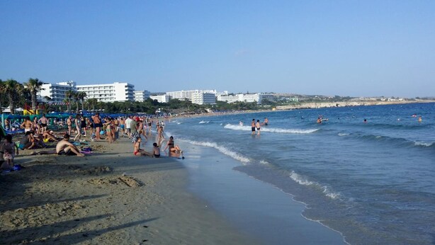 A crowded beach with people sunbathing and swimming, and buildings in the background. - Kkaras Hotel (Ayia Napa)