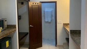 Un pasillo estrecho con una puerta de madera que da a un baño. Se ve un microondas a la izquierda y un bote de basura a la derecha.
