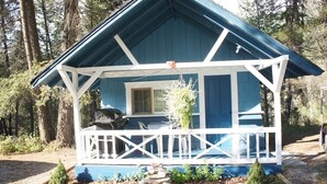 A small, single-story cabin with a blue exterior and white trim, a porch with a railing, and a small garden area in front. - Fairmont Mountain Bungalows (Fairmont Hot Springs)