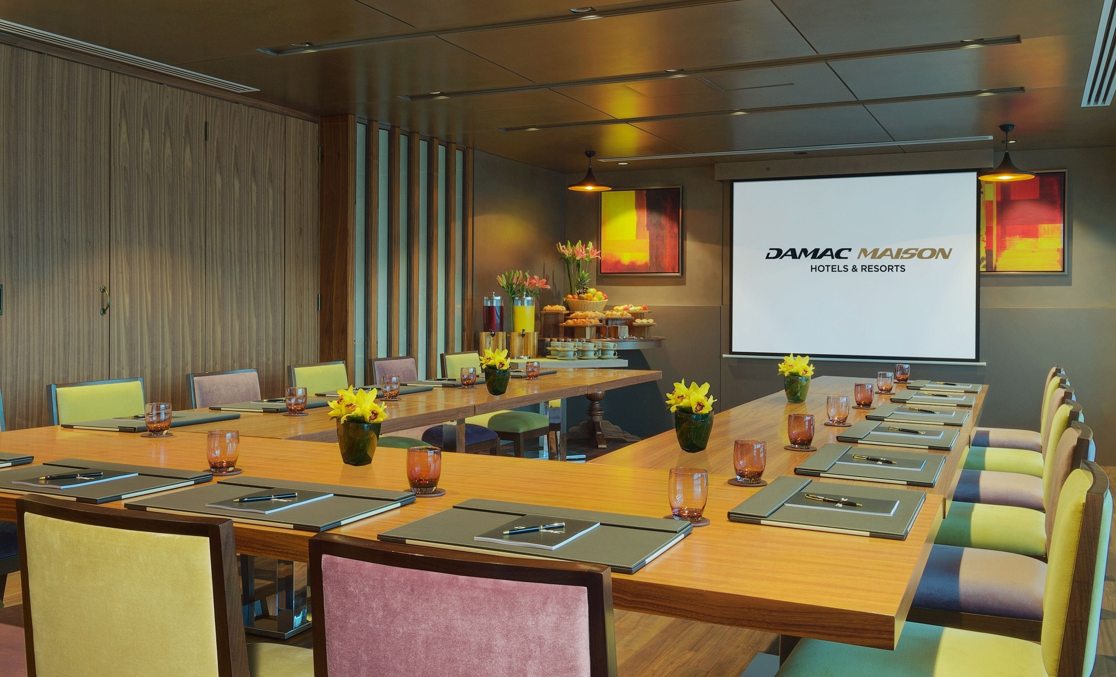 A conference room with a long table, chairs, a projector screen, and a wall with a large logo.
