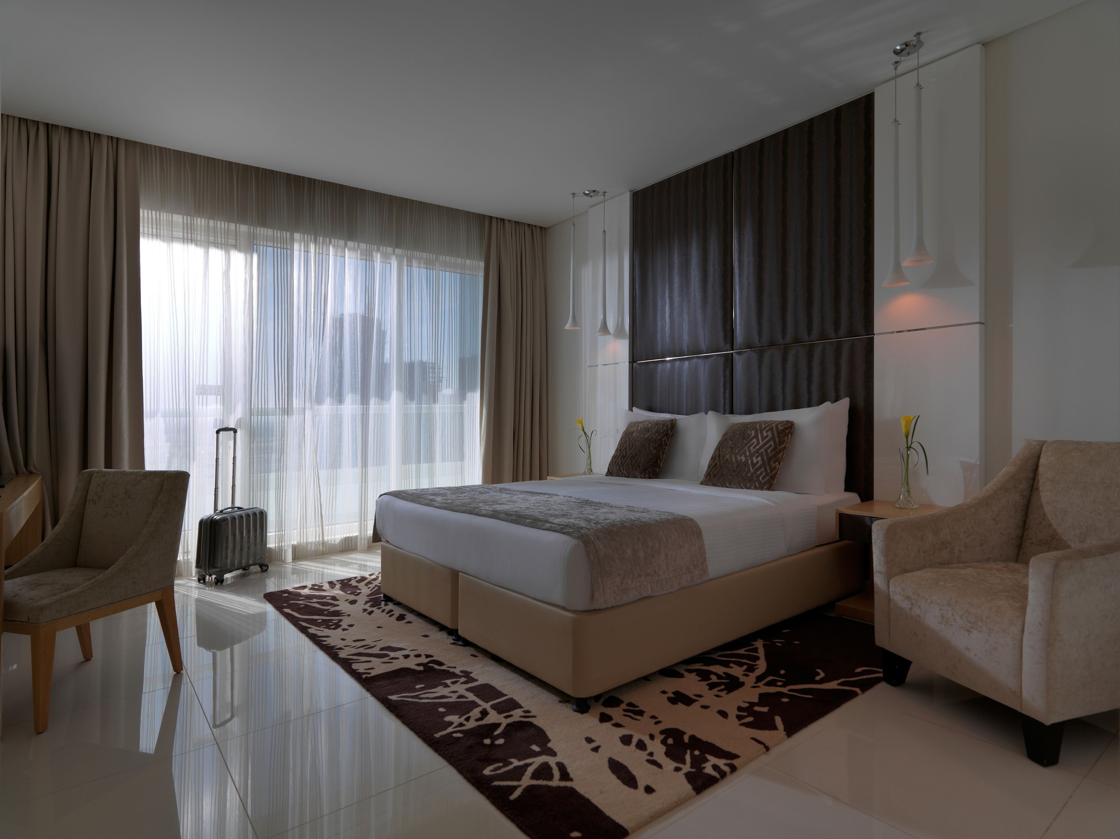 A modern hotel room with a large bed, two armchairs, a small table, a suitcase, and a view of the city through sheer curtains.