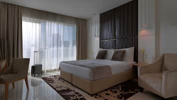 A modern hotel room with a large bed, two armchairs, a small table, a suitcase, and a view of the city through sheer curtains.
