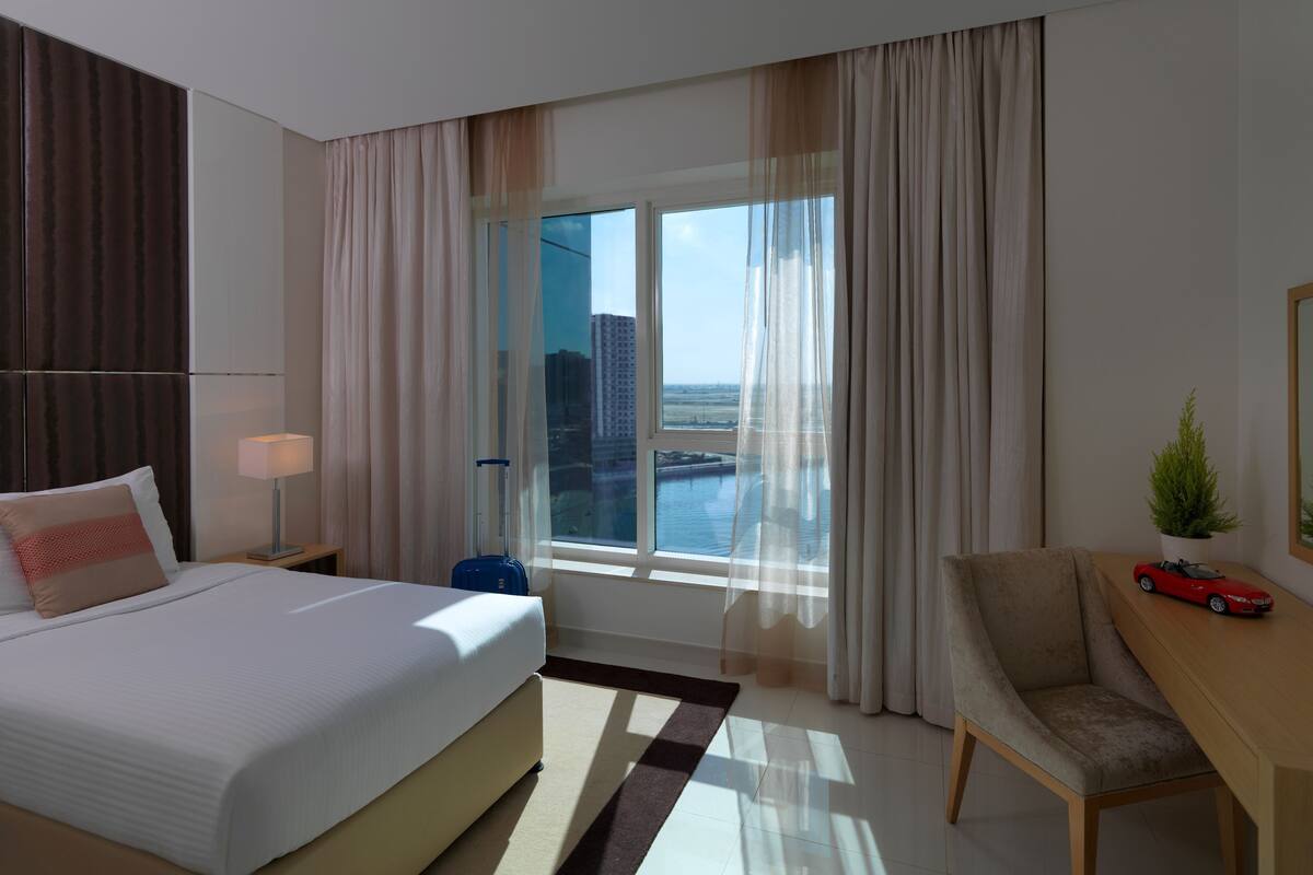 Une chambre d’hôtel avec un grand lit, un bureau avec une chaise, une petite table avec une voiture jouet, et une vue sur le paysage urbain par la fenêtre.