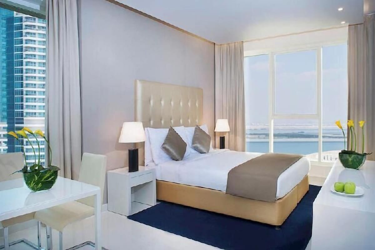 A modern hotel room with a large bed, two bedside lamps, a desk, and a view of the cityscape.