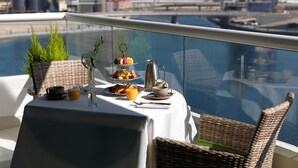 Une table dressée avec un présentoir à trois niveaux pour les desserts, une cafetière et un verre de jus d’orange, sur un balcon donnant sur un paysage urbain et une rivière.