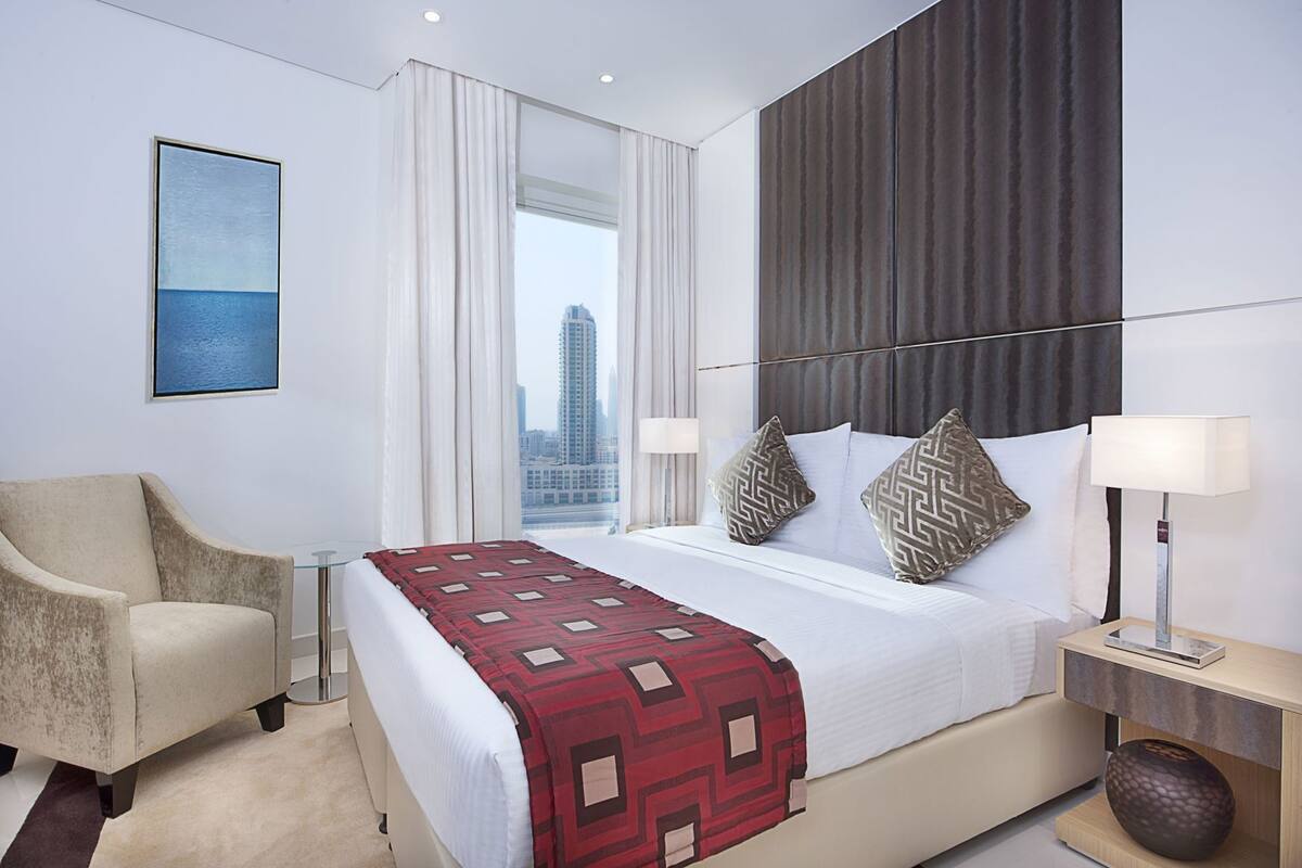 Une chambre d’hôtel avec un lit, une chaise, une table de chevet, une lampe, un vase et une vue sur la ville.