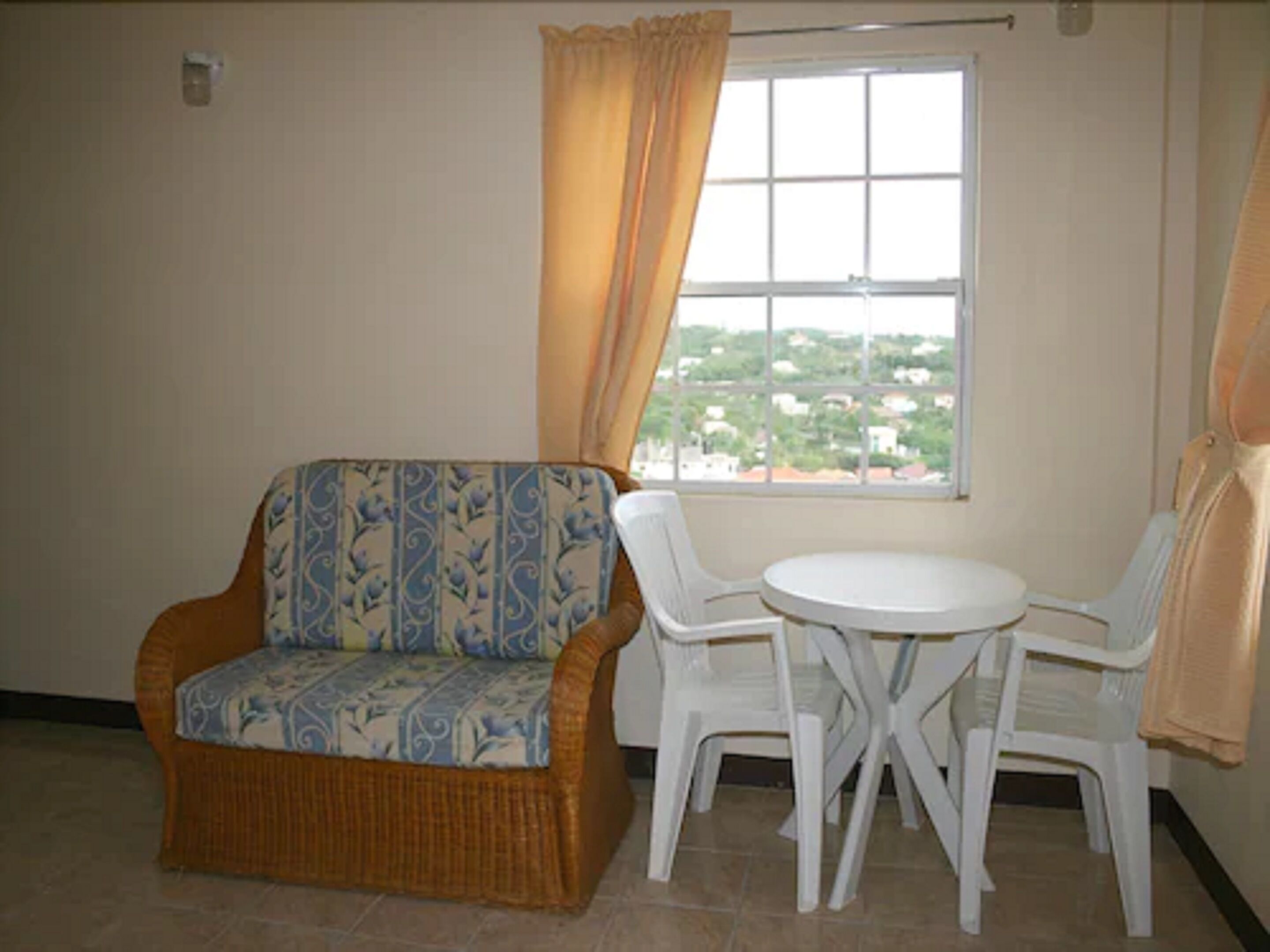 A room with a wicker sofa, two white plastic chairs, a small round table, and a window with a view of buildings.