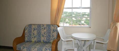 A room with a wicker sofa, two white plastic chairs, a small round table, and a window with a view of buildings.