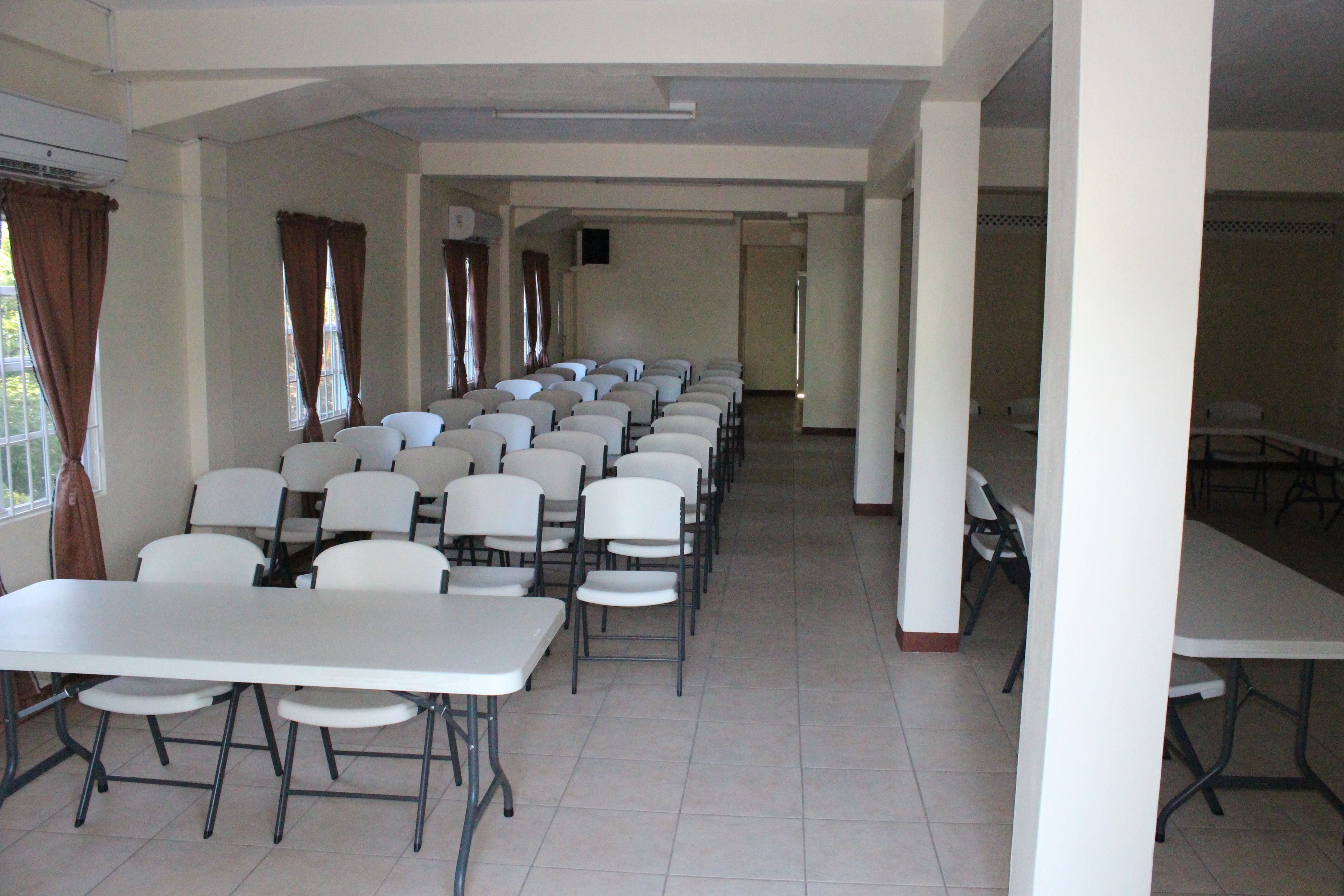 Un salón de conferencias con filas de mesas y sillas blancas, ventanas amplias con cortinas y piso de baldosas.