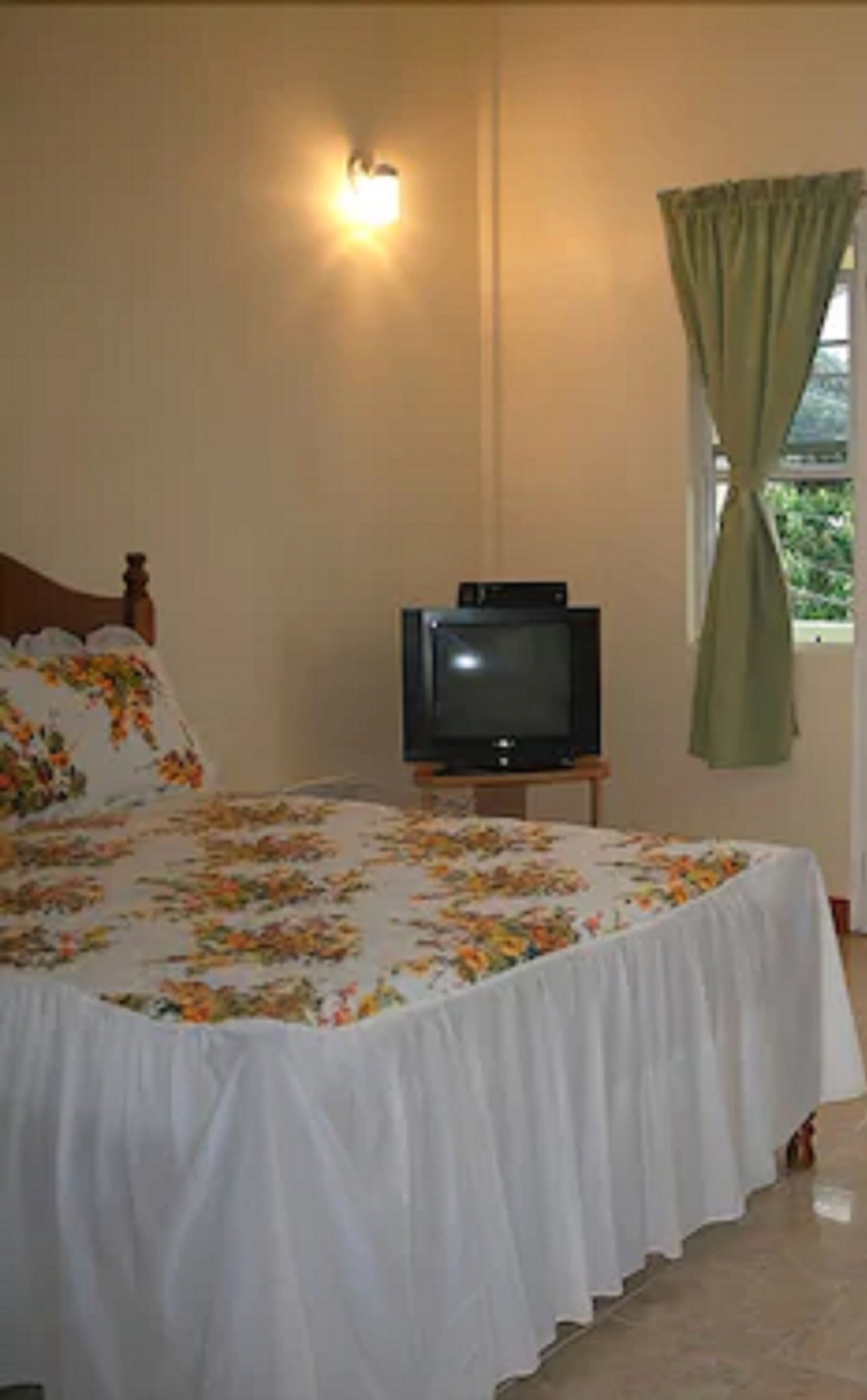 Un dormitorio con una cama, un televisor sobre un soporte, una lámpara de pared y un ventanal con cortinas.
