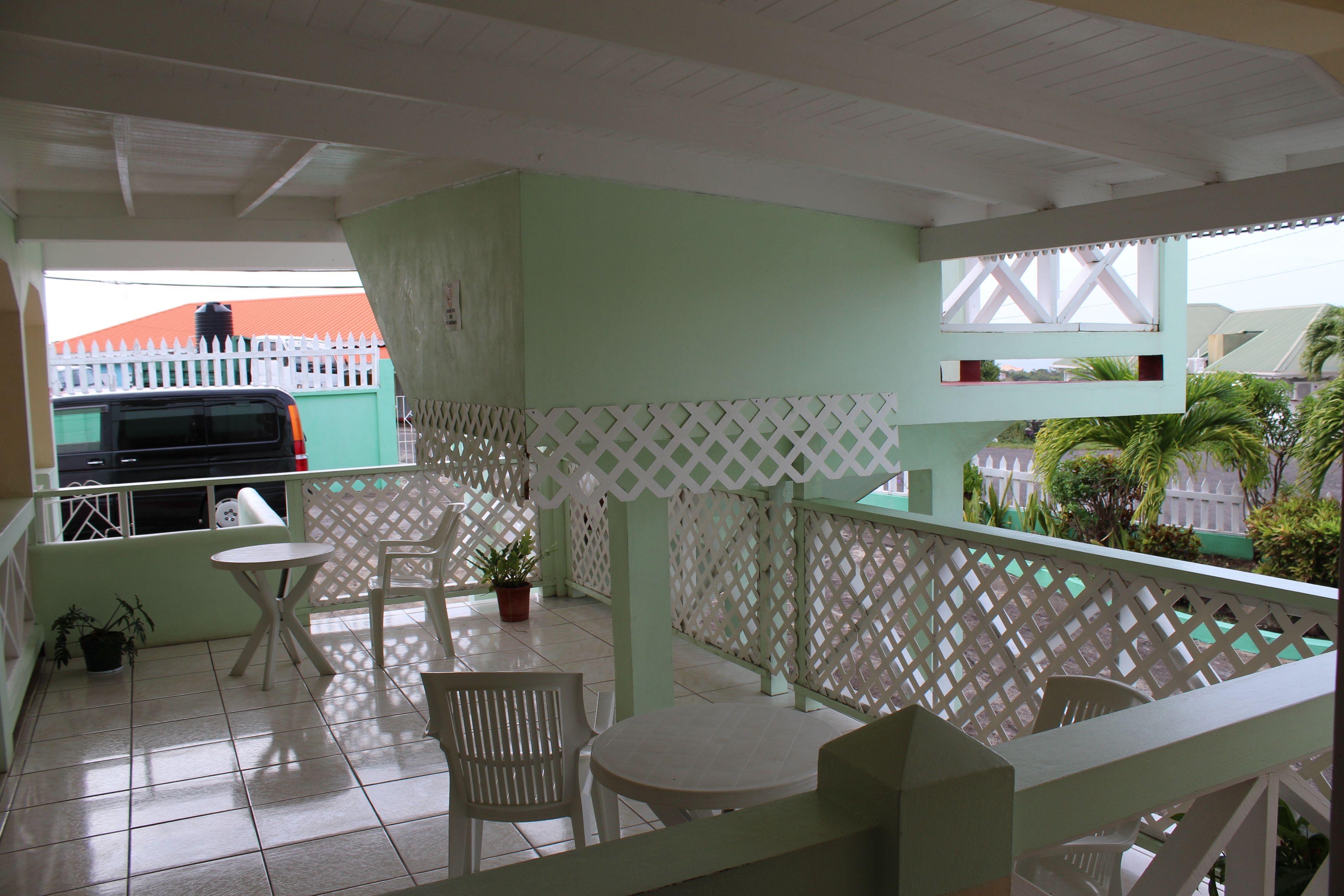 A balcony with white chairs and a table, a grill in the background, and a view of a building with a red roof.