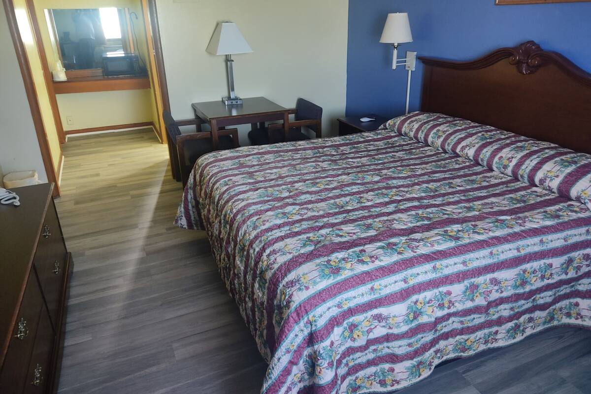 Quarto de hotel com cama, escrivaninha, cadeira, luminária e cômoda.