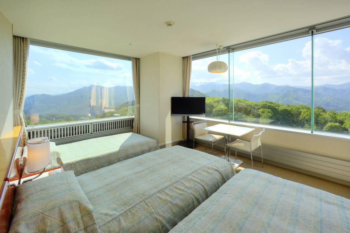 A hotel room with two beds, a desk, a TV, and a large window offering a mountain view.