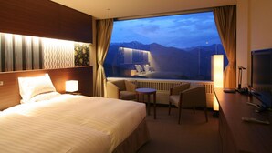 Un cuarto de hotel con una cama grande, un escritorio con computadora, una silla y vistas a las montañas a través de un ventanal amplio.