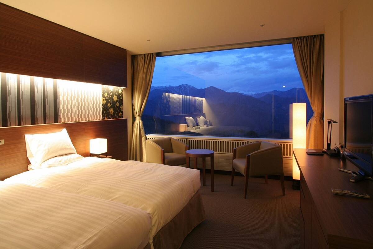 A hotel room with a large bed, a desk with a computer, a chair, and a view of mountains through a large window.
