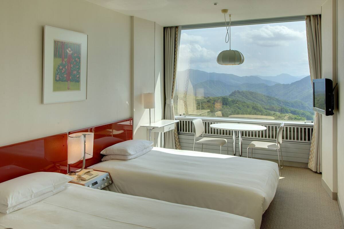 A hotel room with two beds, a desk, a chair, a TV, and a balcony with a mountain view.