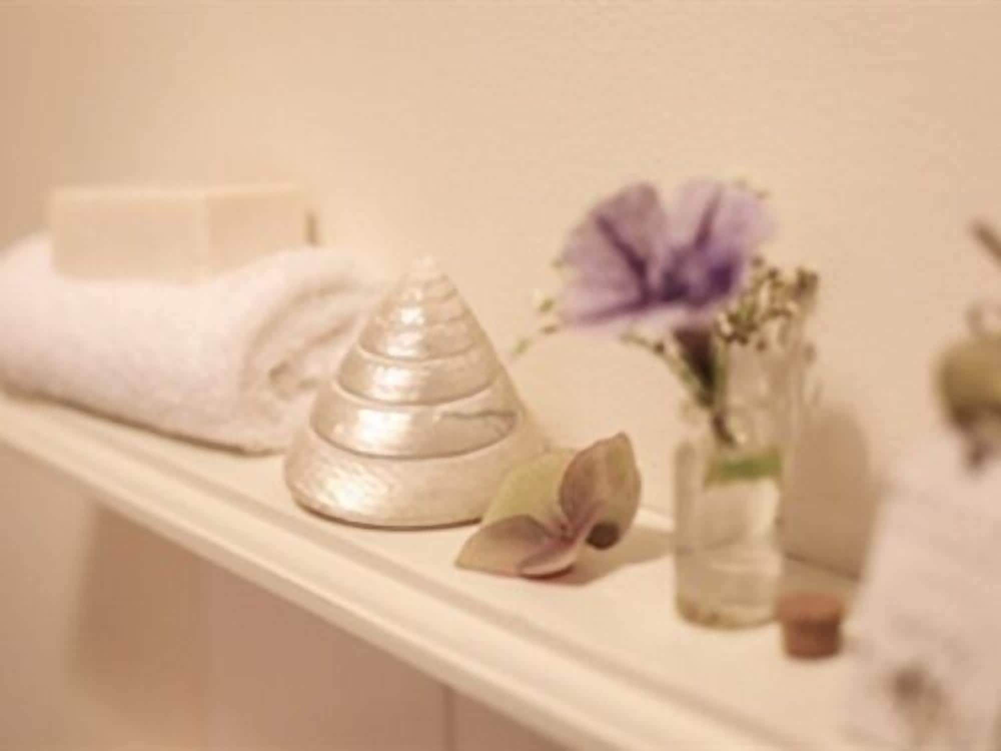 A bathroom shelf with a folded white towel, a silver decorative object, a purple flower, and a small vase with dried flowers.