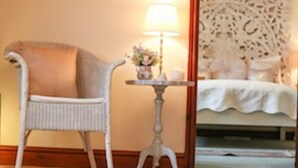 A room with a white upholstered chair, a small round table with a lamp, and a bed with a patterned headboard.