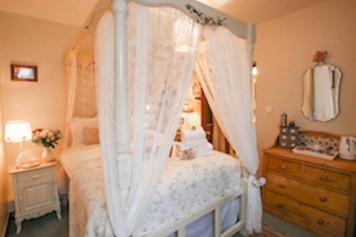 A bedroom with a four-poster bed, a nightstand with a lamp, a dresser, and a mirror on the wall.