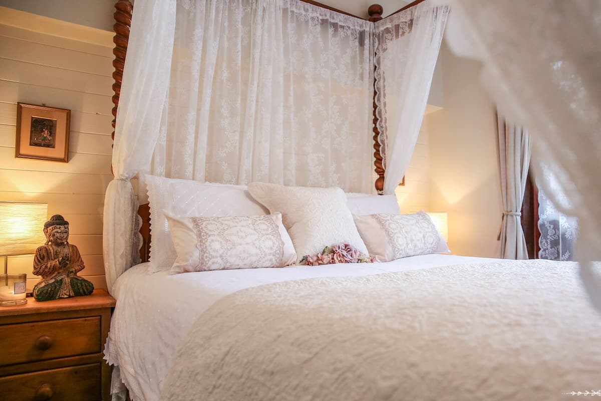 A bedroom with a canopy bed, white bedding, and a bedside table with a lamp and a Buddha statue.