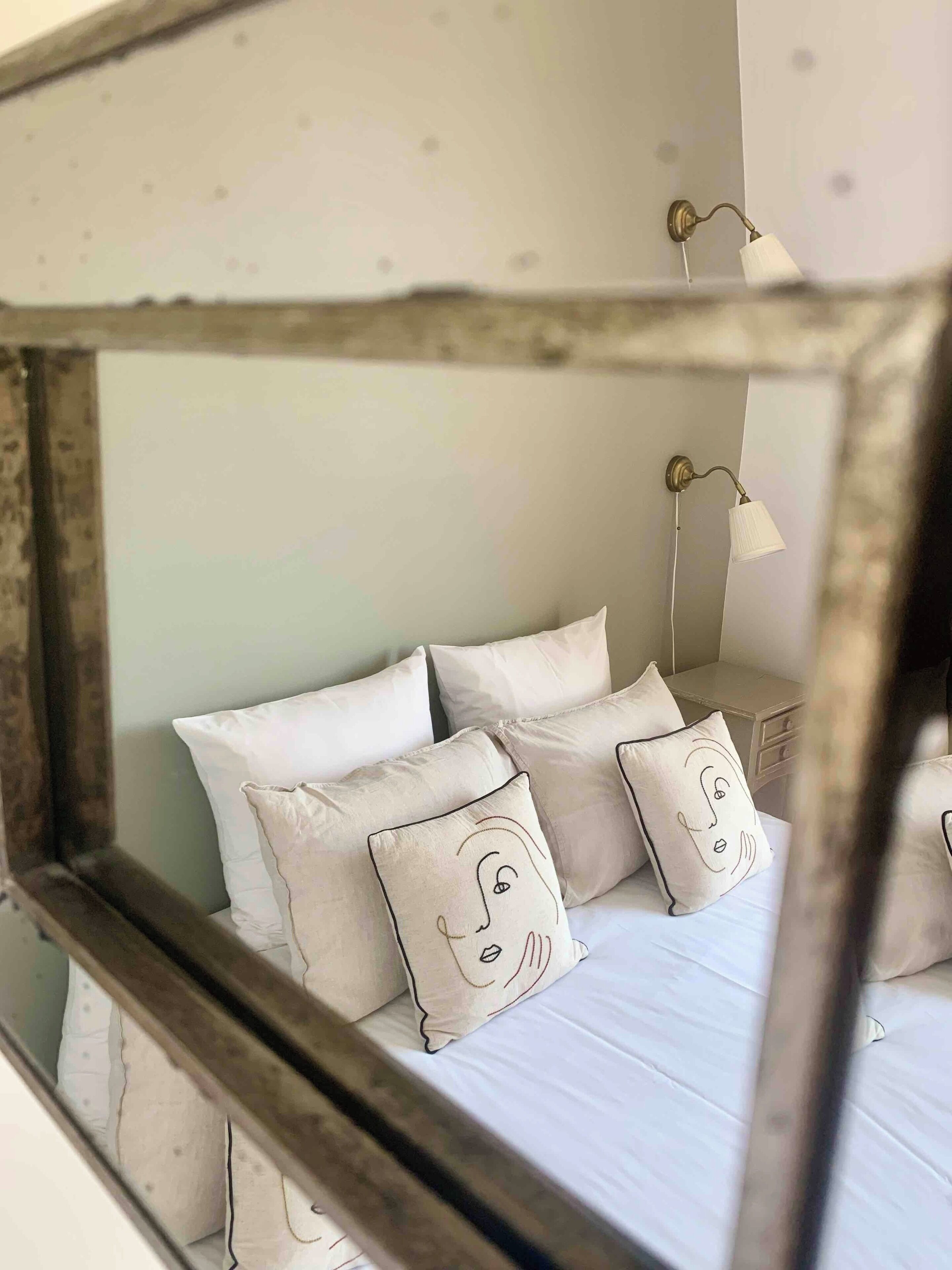 A bed with white bedding and pillows, a nightstand with a lamp, and a mirror reflecting the room.
