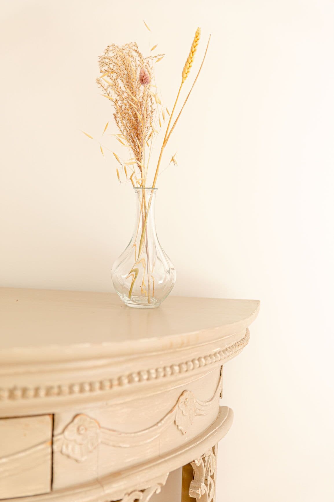 Un florero de vidrio transparente con plantas secas sobre una mesa color crema.