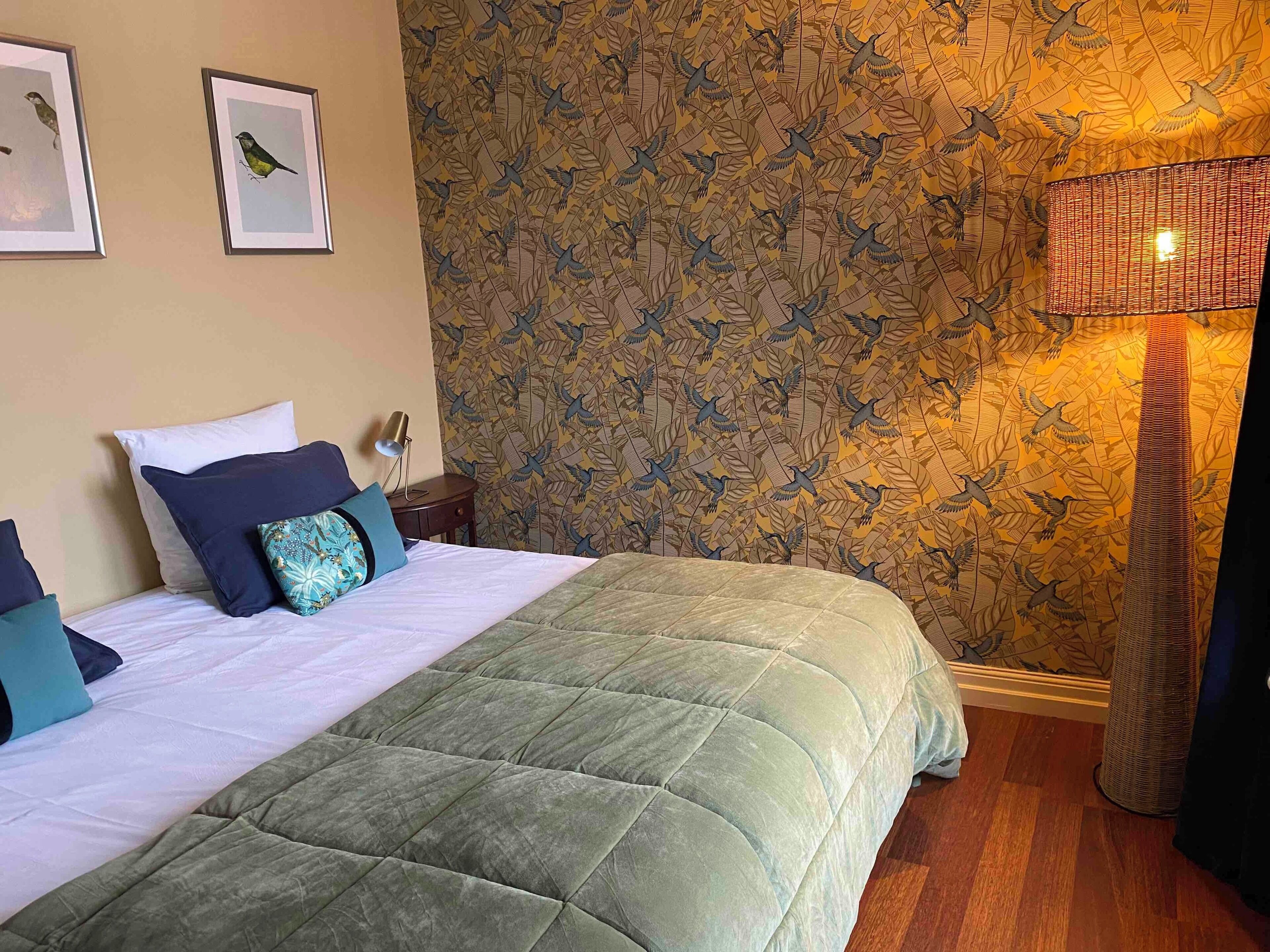 A neatly made bed with a green quilt, a wall with a bird pattern wallpaper, and a lamp on a side table.